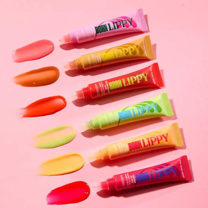 Seven tubes of lip gloss arranged diagonally on a pink background, with corresponding color swatches painted in matching shades below each tube. The tubes are pink, yellow, orange, green, and red, and the brand name 'SOOM LIPPY' is visible on the packaging.