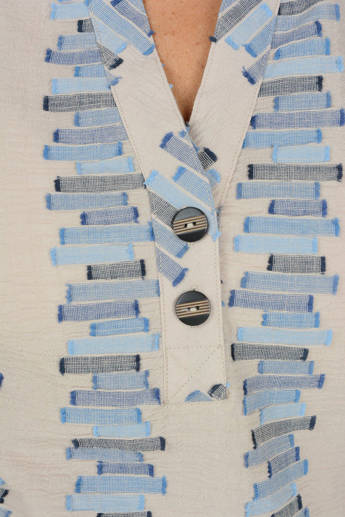 Close-up of a cream-colored textured top with a V-neckline and a decorative button placket. The fabric features vertical rows of light blue, medium blue, and navy striped rectangular patterns.