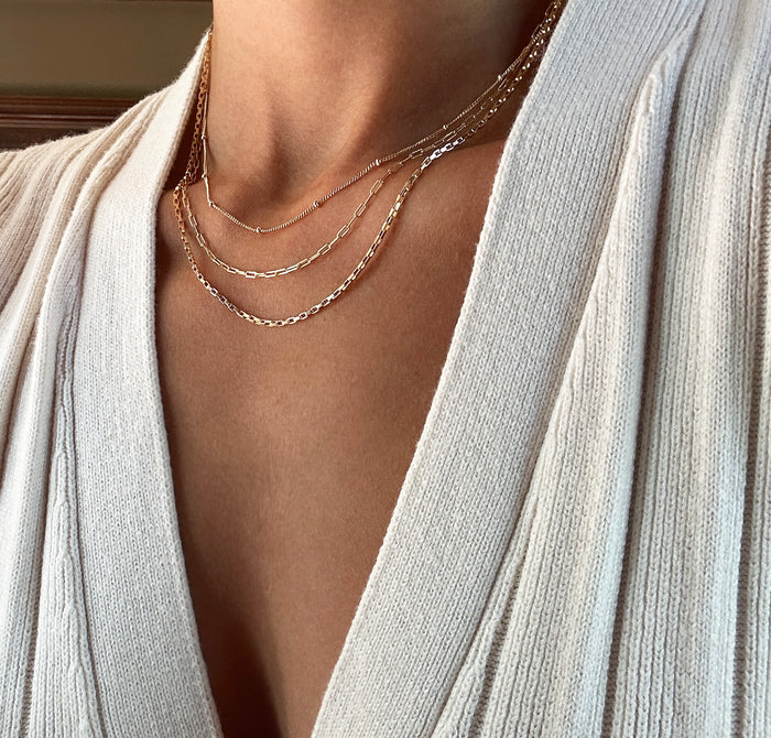 A person wears a cream ribbed V-neck sweater and the LLUME Jewelry Essentials Set—three delicate gold chains of varying lengths, including a paperclip chain.