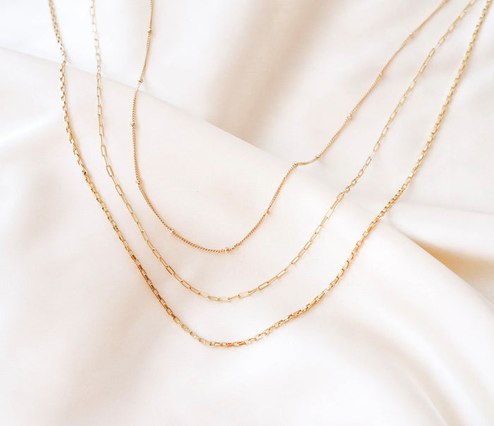 The Essentials Set by LLUME Jewelry features four gold layered necklaces with different link styles, including a paperclip chain, displayed parallel on soft cream satin fabric.