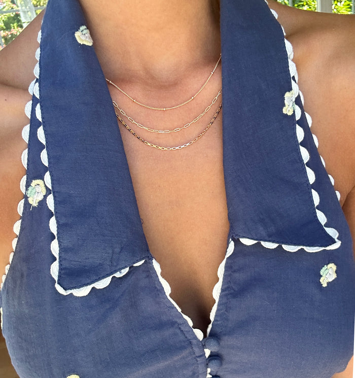A person wearing a navy blue halter dress with scalloped white edges and small embroidered flowers accessorizes with LLUME Jewelry’s Essentials Set, a layered gold necklace set featuring delicate chains and a trendy paperclip design.
