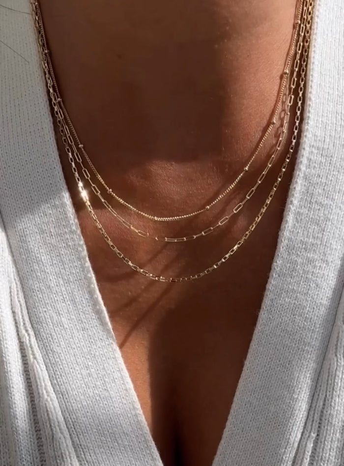 A close-up of a person wearing LLUME Jewelry's Essentials Set: four delicate, layered gold necklaces with varying lengths and styles—including small links and a trendy paperclip chain—paired with a white V-neck top.