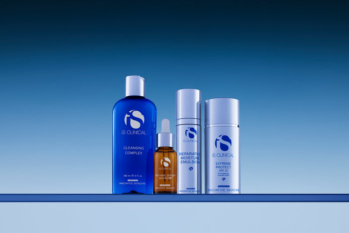 Four iS Clinical skincare products including Cleansing Complex, Pro-Heal Serum Advance+, Reparative Moisture Emulsion, and Extreme Protect SPF 30 on blue surface