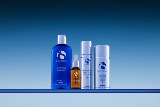 Four iS Clinical skincare products including Cleansing Complex, Pro-Heal Serum Advance+, Reparative Moisture Emulsion, and Extreme Protect SPF 30 on blue surface