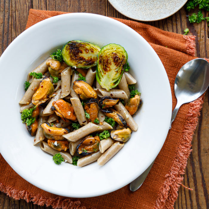 A white bowl of penne pasta with Patagonia Provisions Mussels Lemon Herb, roasted Brussels sprouts, fresh parsley, and a drizzle of Organic Extra Virgin Olive Oil sits on an orange napkin with a spoon on a wooden table.