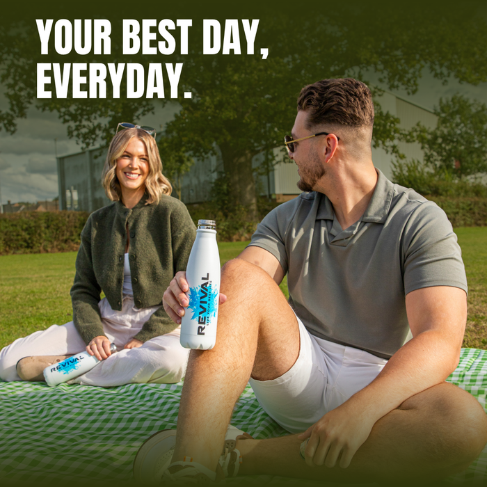 Two people sitting on green checked blanket outdoors holding Revival electrolyte drink bottles, text reads "Your Best Day, Everyday."