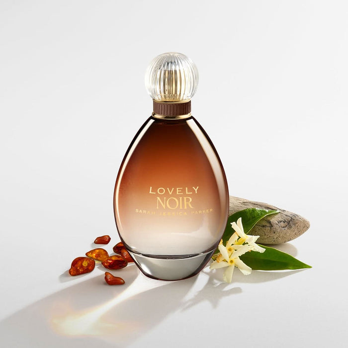 Sarah Jessica Parker Lovely Noir EDP bottle in amber glass with ribbed cap, white flower, green leaves, red stones and rock on white background.
