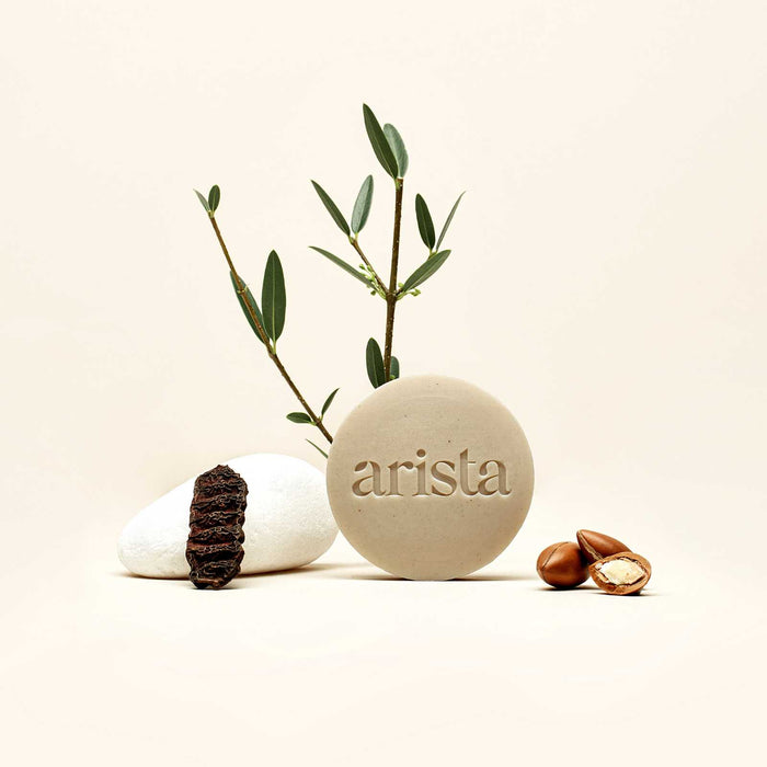 Beige round shampoo bar with arista branding, surrounded by olive branches and smooth stones