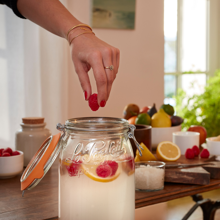 A hand with red-painted nails adds a raspberry to a Le Parfait Storage and Canning Glass Jar, 3L filled with lemon-infused drink on a wooden table, surrounded by raspberries, lemon slices, and assorted fruits epitomizing sustainable canning options.